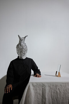 Vertical medium long shot of modern unrecognizable young African American woman wearing black outfit with odd aluminum foil mask sitting at table with mirror on it