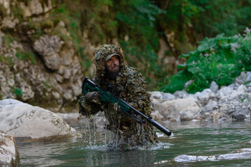 A military man or airsoft player in a camouflage suit sneaking the river and aims from a sniper rifle to the side or to target.