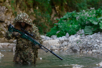 A military man or airsoft player in a camouflage suit sneaking the river and aims from a sniper rifle to the side or to target.