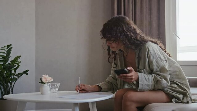 Slow motion of young Caucasian woman with long curly hair sitting in living rom at home writing wedding invitation letter
