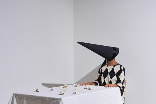 Horizontal conceptual shot of modern unrecognizable African American woman wearing black and white outfit with conus-shaped mask on face sitting at table with cotton flowers on it