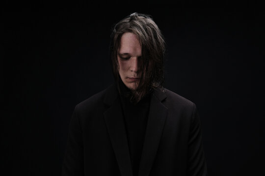 Horizontal medium studio portrait shot of cheerless handsome Caucasian man with long hair wearing black outfit standing in darkness