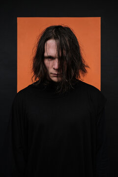 Vertical medium studio portrait of stylish young Caucasian man with messy hairstyle wearing dark clothes standing against orange and black background looking at camera