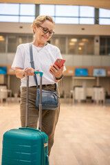 Cheerful elderly woman in glasses with a green suitcase in the lobby of the airport with a...