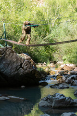 A military man or airsoft player in a camouflage suit sneaking the rope bridge and aims from a sniper rifle to the side or to target. 