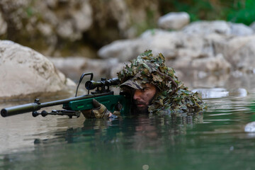 A military man or airsoft player in a camouflage suit sneaking the river and aims from a sniper...