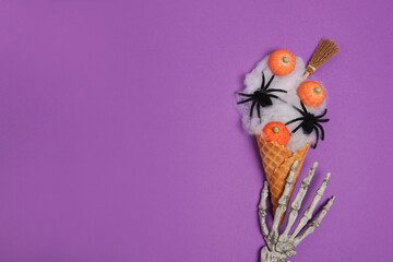 Skeleton hand holding Halloween icecream made with spider web, witch's broom and candies pumpkins on purple background. Halloween sweet dessert. Flat lay, copy space