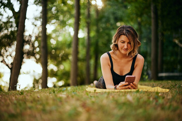 A fit yogi woman is lying on the stomach in forest and using phone.
