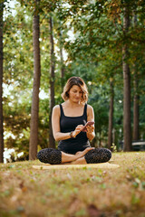 A slim yogi woman sits in a forest in lotus pose and uses phone.