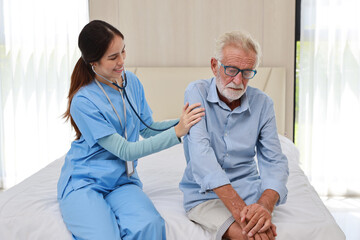 Fototapeta premium Happy caregiver examining senior elderly man patient listening heartbeat using stethoscope&nbsp;on bed at retirement house. Asian smiling nurse taking good help care and support of elder patient at house.