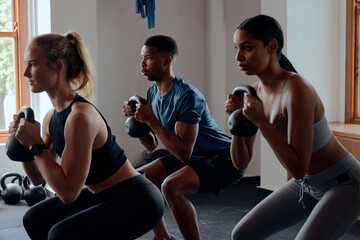 Fototapeta na wymiar Three focused multiracial young adults doing kettlebell squats at the gym
