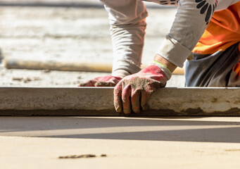 Close up of man builder placing screed rail on the floor covered with sand-cement mix at construction site. Male worker leveling surface with straight edge while screeding floor. Blurred background.
