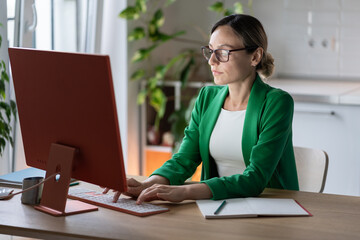 Successful focused Caucasian businesswoman working on computer sits at office desk. Business lady in glasses and green jacket is typing order for company employees or e-mail letter for partners