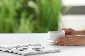 Woman holding cup of coffee at white table indoors, focus on eyeglasses and notebook