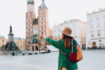 Attractive female tourist is exploring new city. Redhead woman pointing finger and holding a paper map on Market Square in Krakow. Traveling Europe in autumn. St. Marys Basilica. Active lifestyle