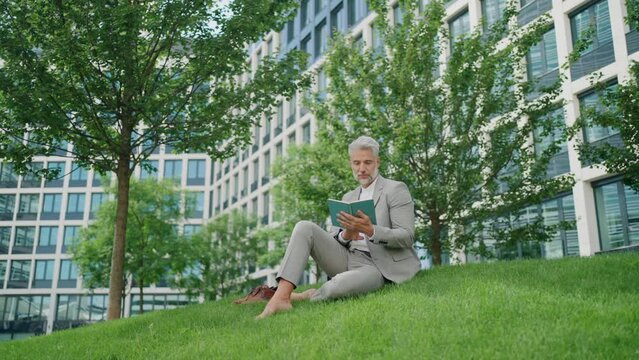 Mature businessman sitting on the grass with a book, in front of office building.