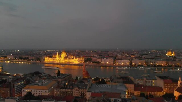 Aerial night seen in front of the illuminated Hungarian Parliament. City sightseeing ships cruise on the Danube. Pest and Buda on the two river banks.