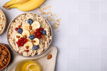 Bowl of oatmeal berries, almonds and banana slices on white tiled table, flat lay. Space for text