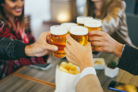Group of people having fun cheering with beer inside brewery bar - Focus on front hand holding glass