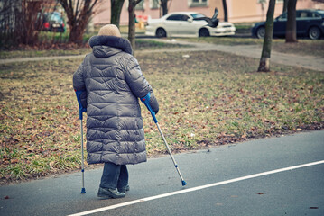 Fototapeta premium Old woman walk on the street with forearm crutches in hands. Оld female on canadian crutches walking on city street. Senior walking with crutches, orthopedic problems, joint disorder.
