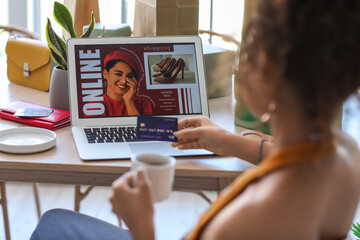 Young African-American woman with cup of coffee and credit card shopping online at home