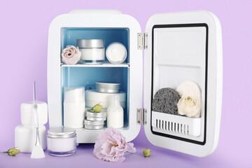 Small refrigerator with cosmetics and flowers on lilac background