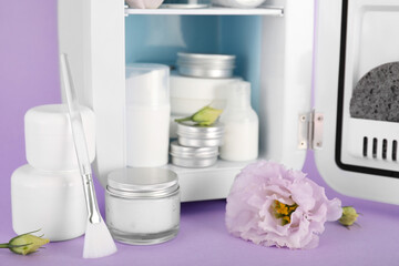 Small refrigerator with cosmetics and flowers on lilac background, closeup