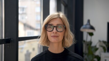 Blonde woman portrait in eyeglasses with tense emotions on background of office room. Attractive stylish person looking at the camera with neutral expression. Concept of businesswoman. 
