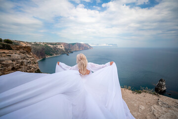Blonde with long hair on a sunny seashore in a white flowing dress, rear view, silk fabric waving in the wind. Against the backdrop of the blue sky and mountains on the seashore.