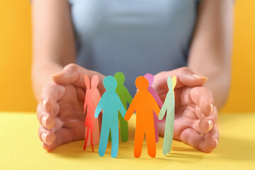 Woman protecting paper human figures on yellow table, closeup. Diversity and inclusion concept