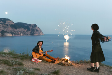 Romantic by the campfire in the mountains with a view of the sea. Man plays the guitar and a woman...