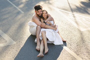 A man and a woman in white blankets are sitting on an empty city road in the morning.