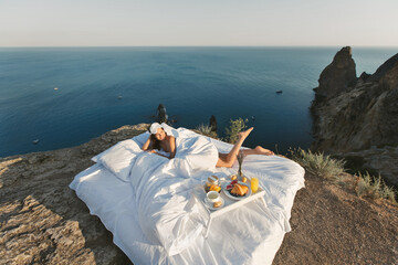A beautiful young woman wakes up alone in a white bed on a cliff overlooking the sea.