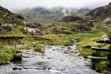 Scenic shot of a trickling river surrounded with green hills on a foggy overcast day