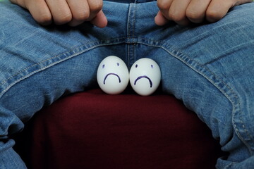 Young Asian man holding two chicken eggs with sad face on crotch groin area. Male infertility, testicular cancer and impotence concept.  - obrazy, fototapety, plakaty