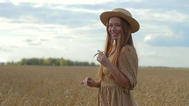 Portrait of an attractive young woman in summer dress and hat standing on wheat field against beautiful sunset looking into camera and laughing. happy person is resting outside in  field