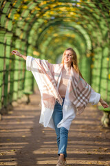 Naklejka premium Beautiful happy young woman with light brown hair walks along a picturesque alley in an autumn park. An attractive woman wrapped herself in a plaid. Fall season. Amazing autumn mood.