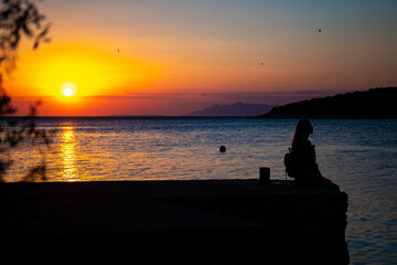 dark silhouette of a beautiful long-haired girl sitting by the seashore in croatia during a colorful sunset; romantic sunset walk by the mediterranean sea