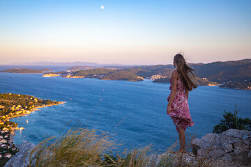 beautiful long-haired girl in a long dress stands on top of a mountain enjoying the sunset over the...