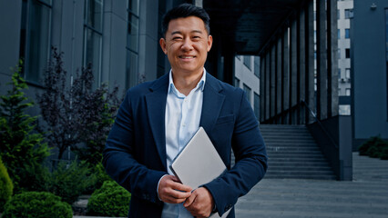Asian 40s middle-aged businessman entrepreneur employer man worker manager designer financial analyst standing in city near office company building in downtown posing holding laptop looking at camera - obrazy, fototapety, plakaty