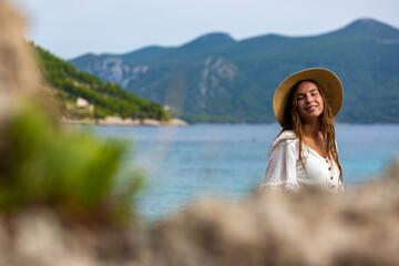 beautiful tanned woman in white dress and hat rests on the rocks during sunset over paradise beach...