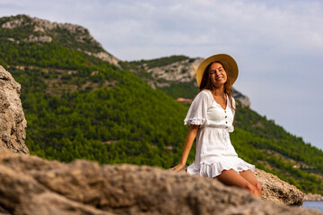beautiful tanned woman in white dress and hat rests on the rocks during sunset over paradise beach in croatia with green mountains in the background; peljesac peninsula 