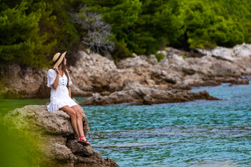beautiful tanned woman in white dress and hat rests on the rocks during sunset over paradise beach in croatia with green mountains in the background; peljesac peninsula 