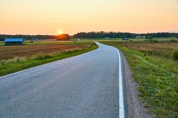 Empty asphalt road surrounded by fields and trees during the sunset