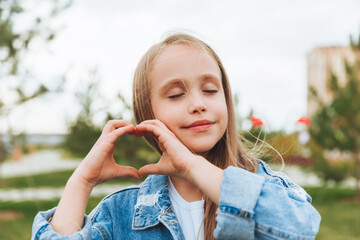 A little blonde girl shows a heart sign made with her fingers while standing on the street in the park