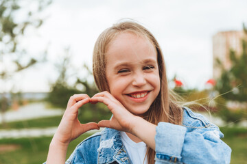 A little blonde girl shows a heart sign made with her fingers while standing on the street in the park