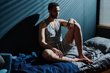 Young sleepless man in underwear sitting on bed in dark room at midnight and suffering from post traumatic syndrome - obrazy, fototapety, plakaty
