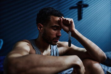 Obraz premium Young unhappy man with post traumatic syndrome holding silver chain with metallic medallion in hand touching his forehead