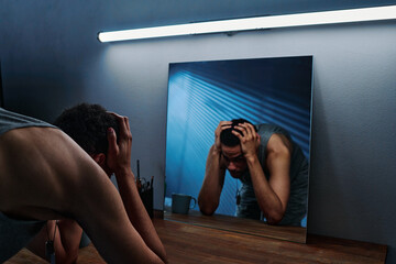 Young man with post traumatic disorder standing in front of mirror with his head in hands while...