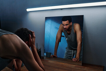 Young insomniac man holding head in hands in front of mirror with reflection of him expressing...